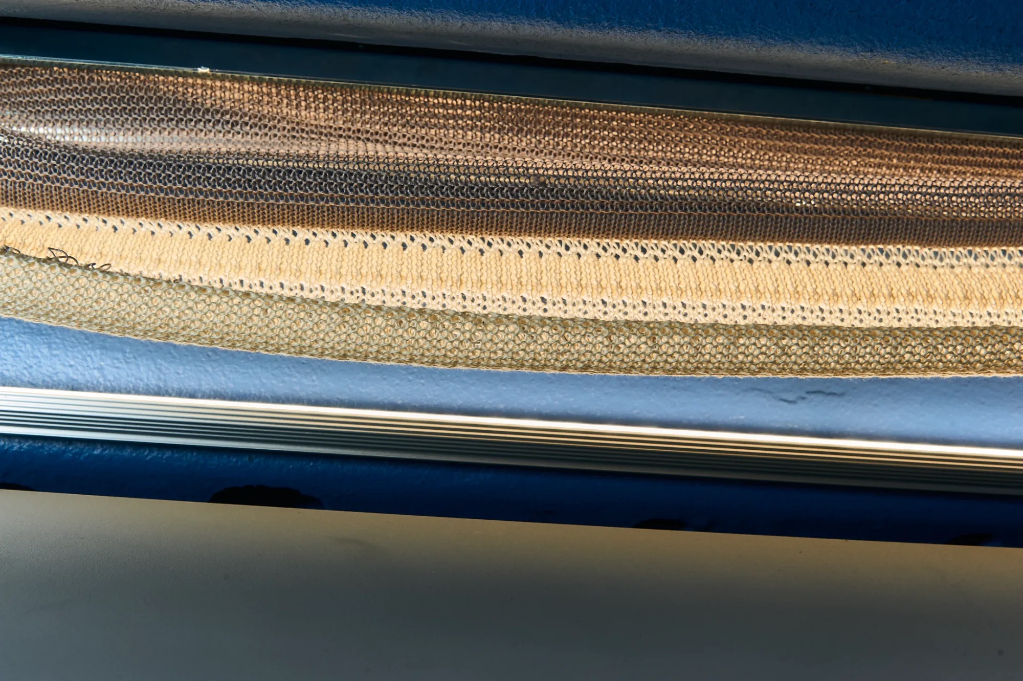 Bands of technical knit in tan, ecru and open mesh structures coming off the bed of a blue flatbed machine